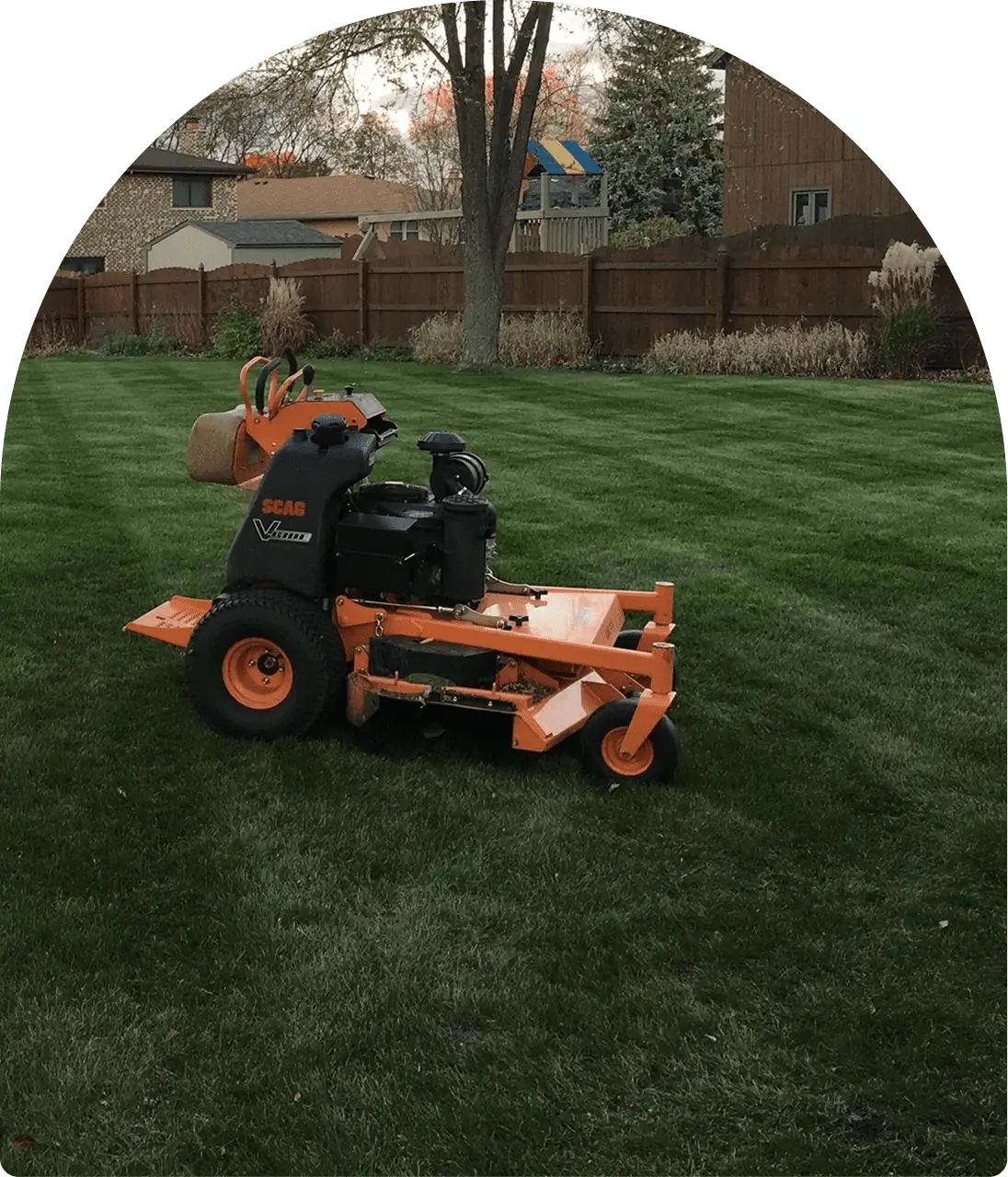 Commercial lawn mower on freshly mowed grass in a backyard with trees, fence, and houses in the background.