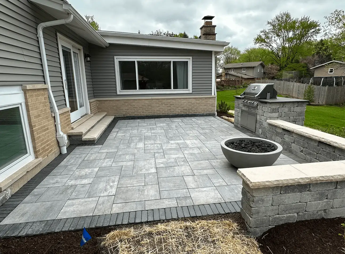 Backyard patio with gray stone pavers, a built-in grill, a round fire pit, and steps leading to a house with beige and gray siding. A green lawn and fence are in the background.